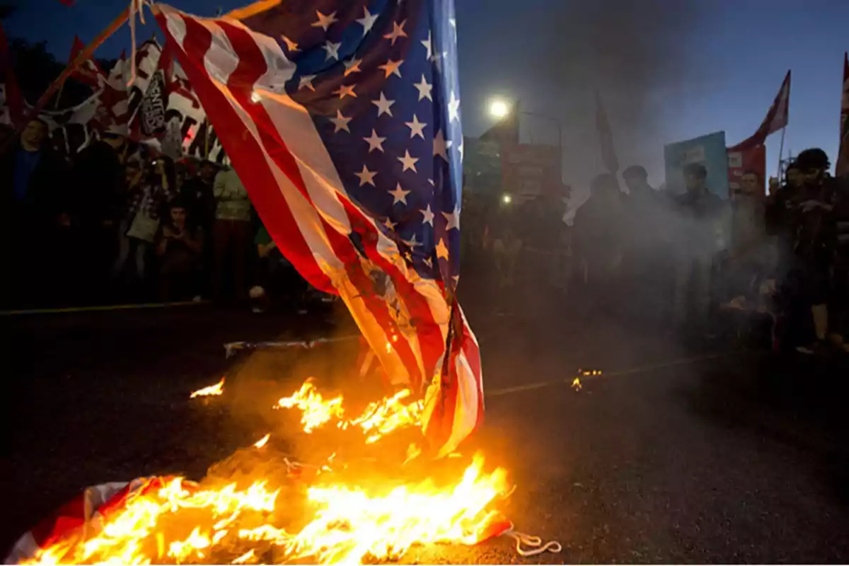 United States flag burning on the ground during a protest with people and signs in the background United States flag burning on the ground during a protest with people and signs in the background