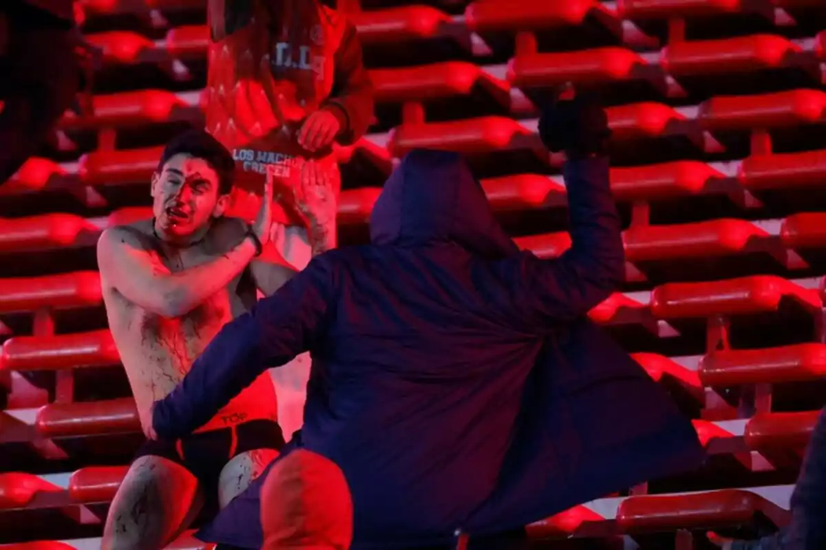 A jerseyless man with blood on his face shields himself from a blow while another person in a dark coat confronts him on stairs lit in red. A jerseyless man with blood on his face shields himself from a blow while another person in a dark coat confronts him on stairs lit in red.