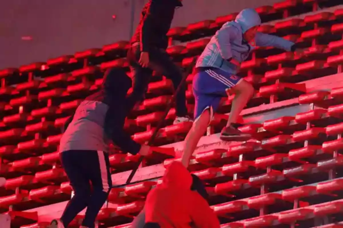 Personas con ropa deportiva y capuchas suben por las gradas de un estadio iluminado con luces rojas