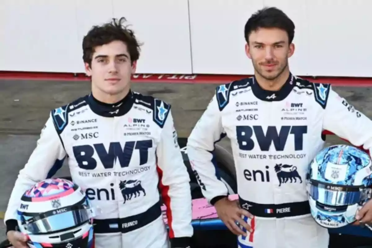Two racing drivers in white BWT Alpine team suits pose with their helmets in front of a pit lane backdrop. Two racing drivers in white BWT Alpine team suits pose with their helmets in front of a pit lane backdrop.