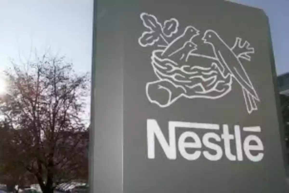 Letrero gris de Nestlé con el logo de un nido y pájaros al aire libre junto a un árbol Letrero gris de Nestlé con el logo de un nido y pájaros al aire libre junto a un árbol