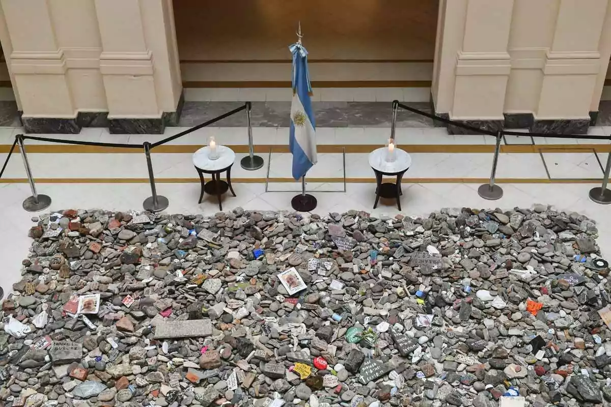 Montón de piedras con mensajes y fotos, rodeado por una cuerda de seguridad, dos mesas con velas encendidas y una bandera de Argentina en el centro, en el interior de un edificio. Montón de piedras con mensajes y fotos, rodeado por una cuerda de seguridad, dos mesas con velas encendidas y una bandera de Argentina en el centro, en el interior de un edificio.