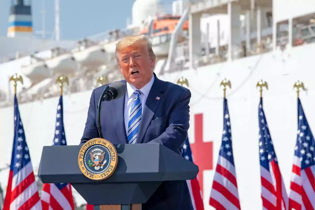 Man in a blue suit speaking at a presidential podium in front of a white ship with United States flags in the background