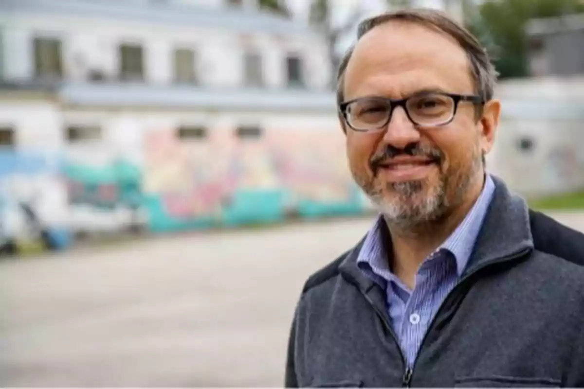 Middle-aged man with glasses and a beard smiles outdoors with a building and a colorful mural blurred in the background Middle-aged man with glasses and a beard smiles outdoors with a building and a colorful mural blurred in the background