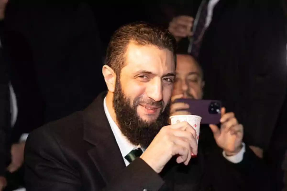 Bearded man in a dark suit smiles while holding a small glass at a social gathering.