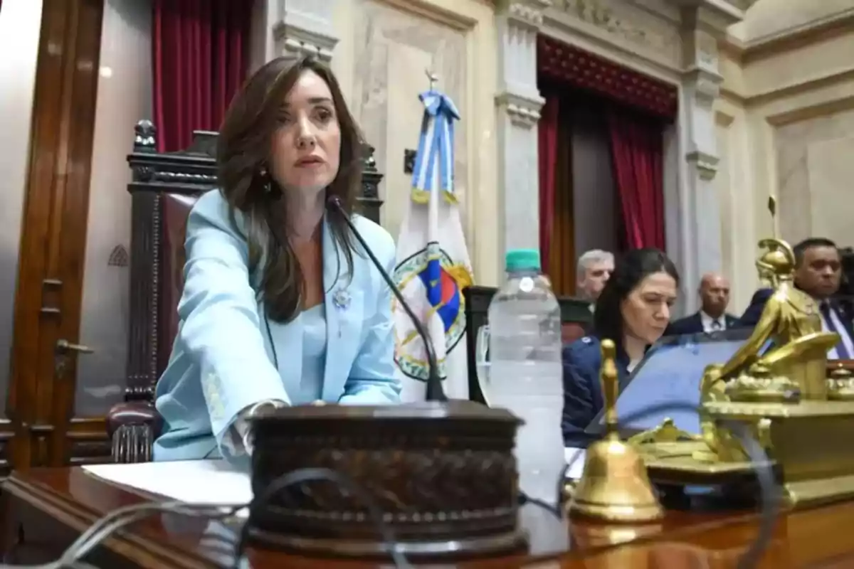 Woman in a light blue blazer seated at a parliamentary podium with a microphone and golden objects on the table; in the background, flags and other people are visible. Woman in a light blue blazer seated at a parliamentary podium with a microphone and golden objects on the table; in the background, flags and other people are visible.