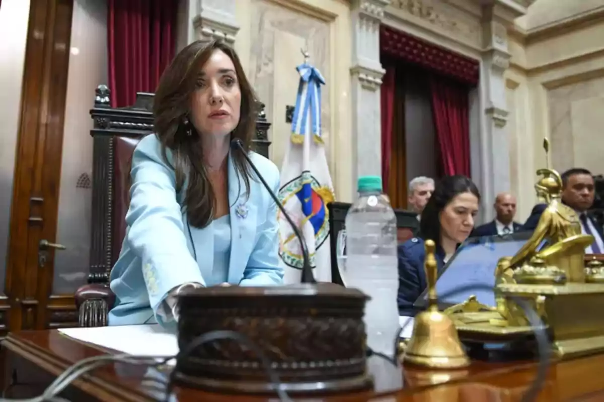 Woman in a light blue blazer seated at a parliamentary podium, surrounded by official objects and people working in the background