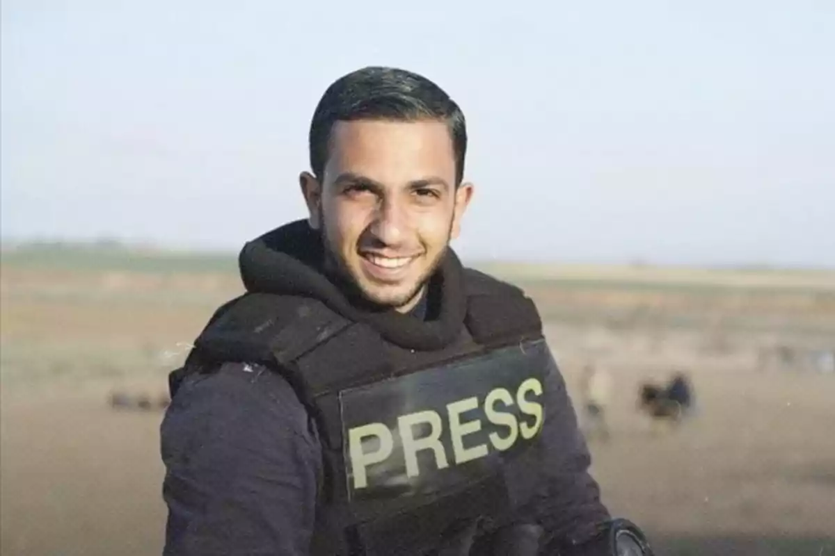 A smiling journalist wearing a bulletproof vest labeled PRESS in an open field