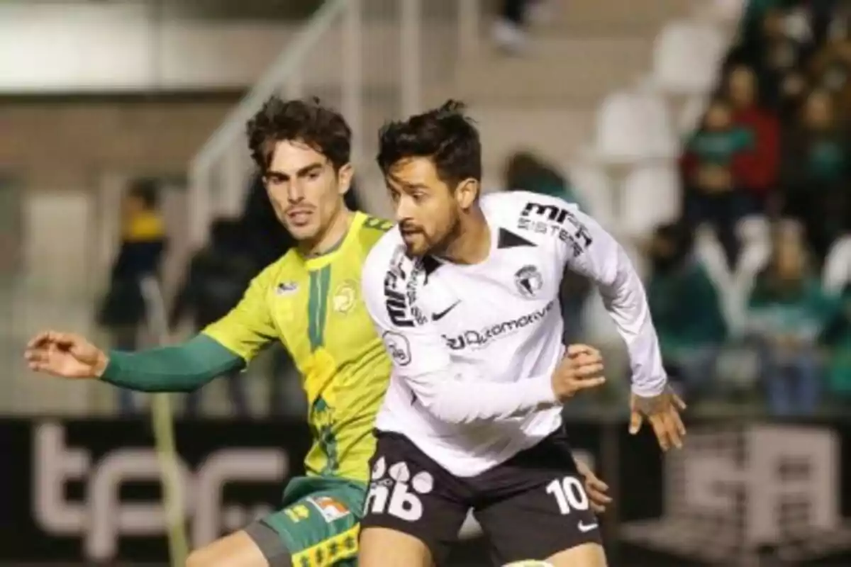 Dos futbolistas disputan el balón durante un partido en un estadio con público en las gradas. Dos futbolistas disputan el balón durante un partido en un estadio con público en las gradas.