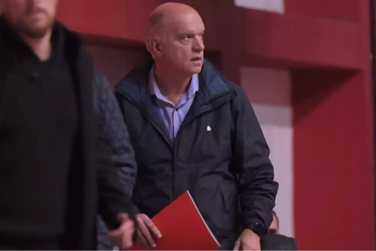 Short-haired, gray-haired man in a dark jacket holding a red folder in an indoor setting with red and white walls Short-haired, gray-haired man in a dark jacket holding a red folder in an indoor setting with red and white walls