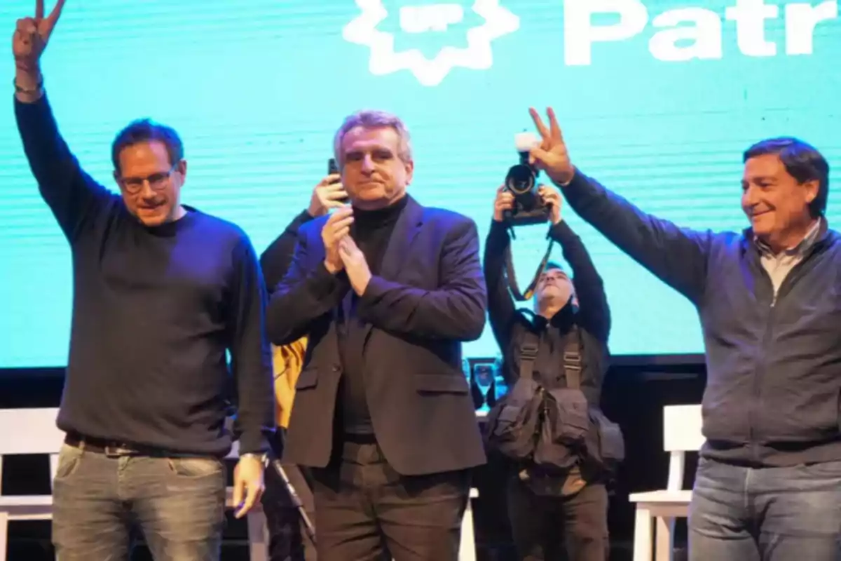 Three men on a stage, two of them raising their hands making the victory sign while a photographer takes pictures in the background. Three men on a stage, two of them raising their hands making the victory sign while a photographer takes pictures in the background.