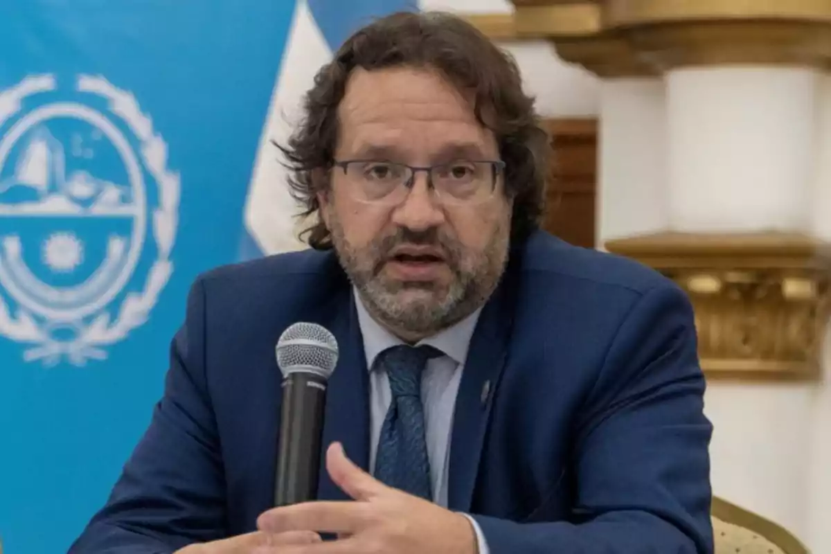 Hombre de cabello rizado y barba con gafas y traje azul hablando en una conferencia mientras sostiene un micrófono Hombre de cabello rizado y barba con gafas y traje azul hablando en una conferencia mientras sostiene un micrófono