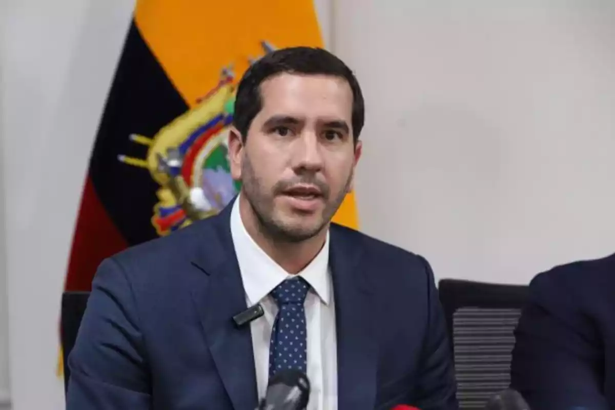 Hombre de cabello corto y barba recortada usando traje azul y corbata, sentado frente a un micrófono con una bandera de Ecuador al fondo