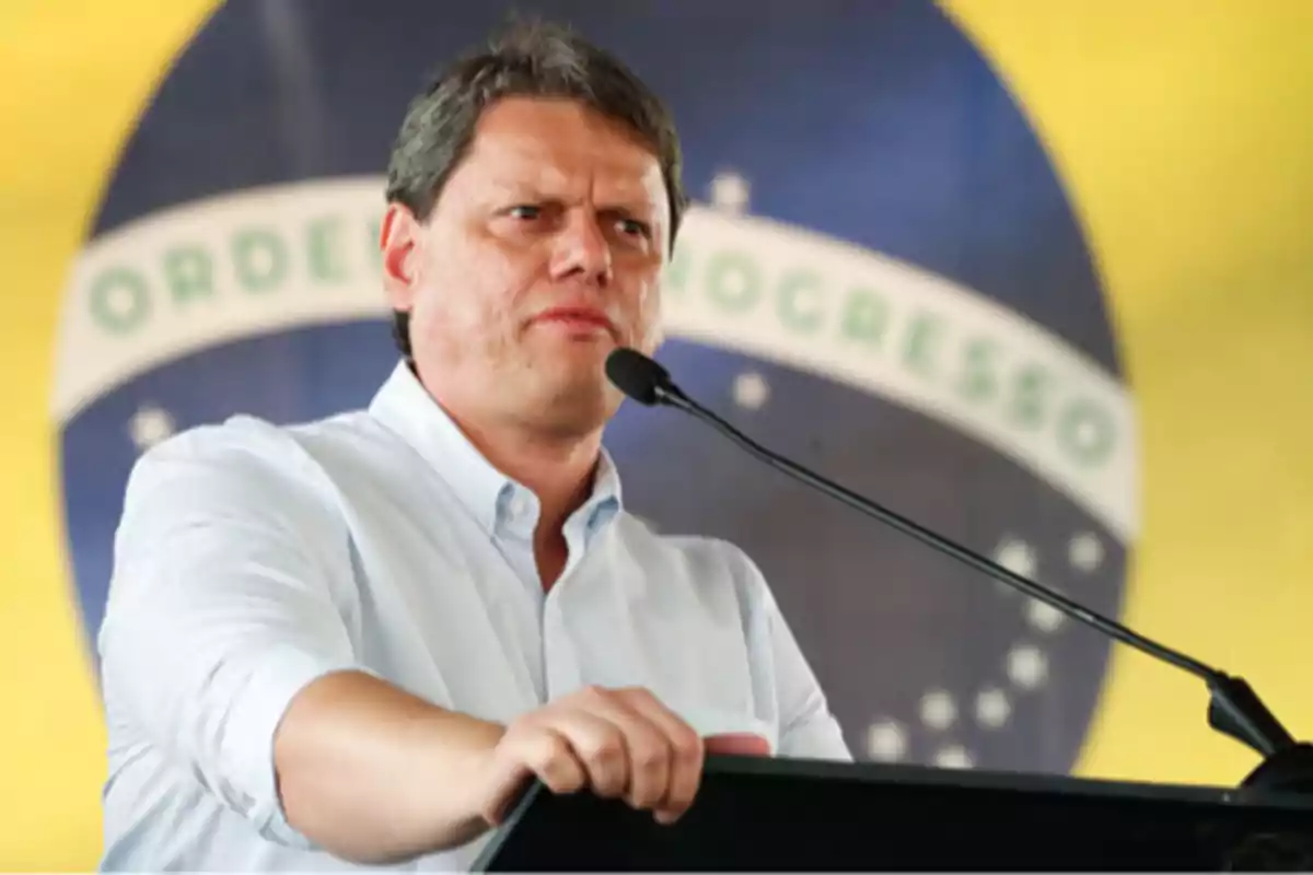 Short-haired man in a light-colored jersey speaking at a podium with a microphone in front of a blurred Brazilian flag in the background Short-haired man in a light-colored jersey speaking at a podium with a microphone in front of a blurred Brazilian flag in the background