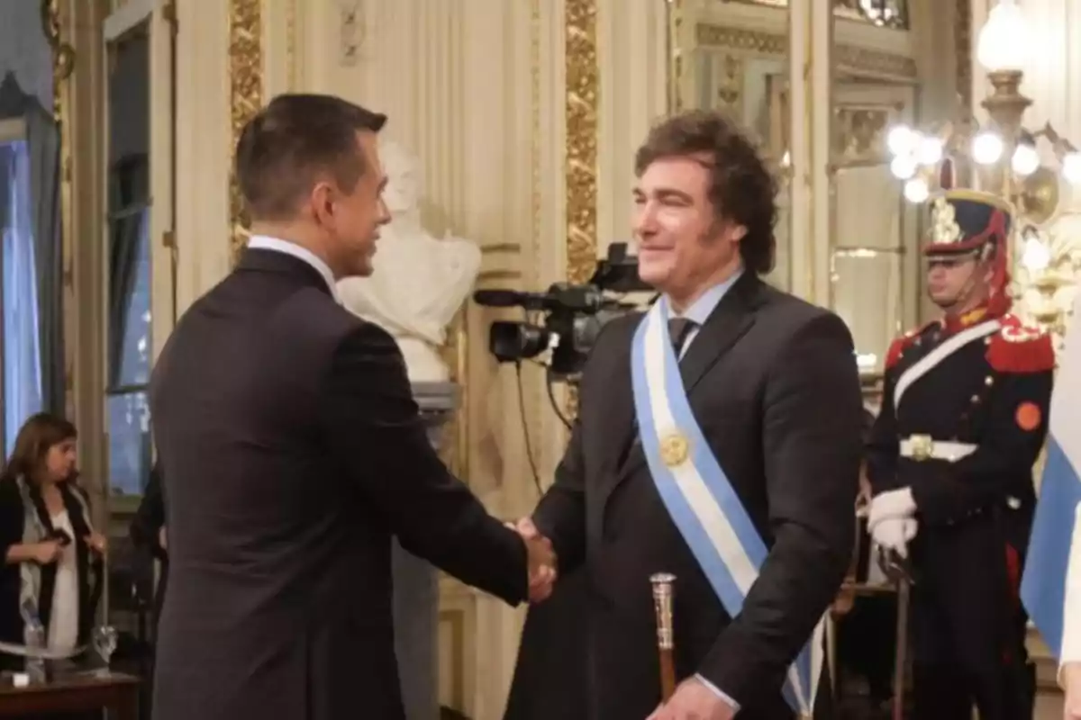 Two men in suits shake hands in an elegant hall; one of them wears a presidential sash and holds a staff while a guard in dress uniform stands in the background.