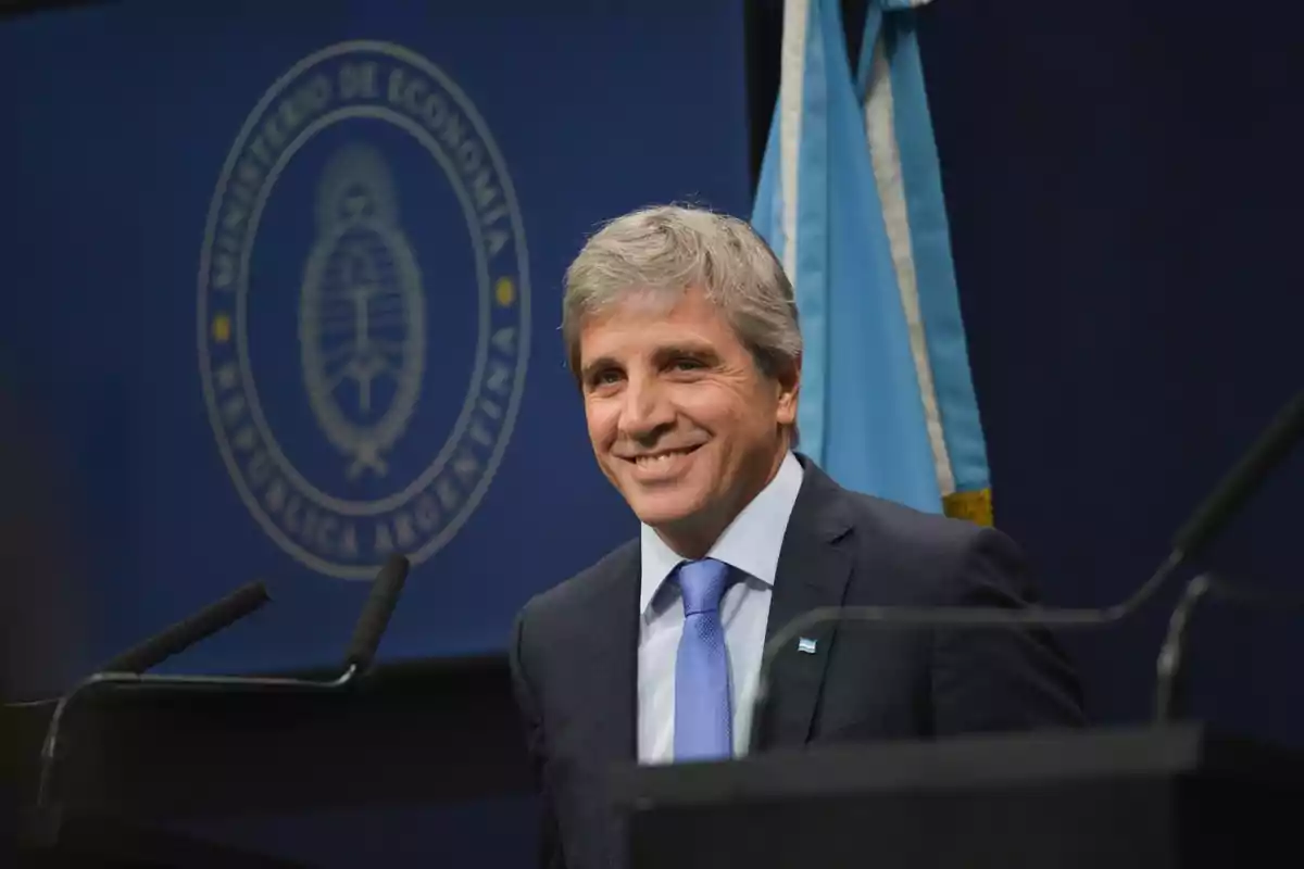 Hombre de cabello canoso con traje y corbata azul sonriendo frente a un atril con la bandera de Argentina y el logo del Ministerio de Economía de fondo Hombre de cabello canoso con traje y corbata azul sonriendo frente a un atril con la bandera de Argentina y el logo del Ministerio de Economía de fondo