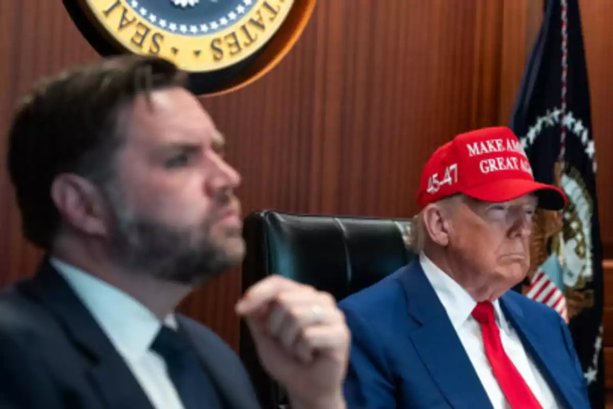 Two men in suits in a meeting room, one of them wearing a red cap with a message and a background featuring the presidential seal of the United States. Two men in suits in a meeting room, one of them wearing a red cap with a message and a background featuring the presidential seal of the United States.