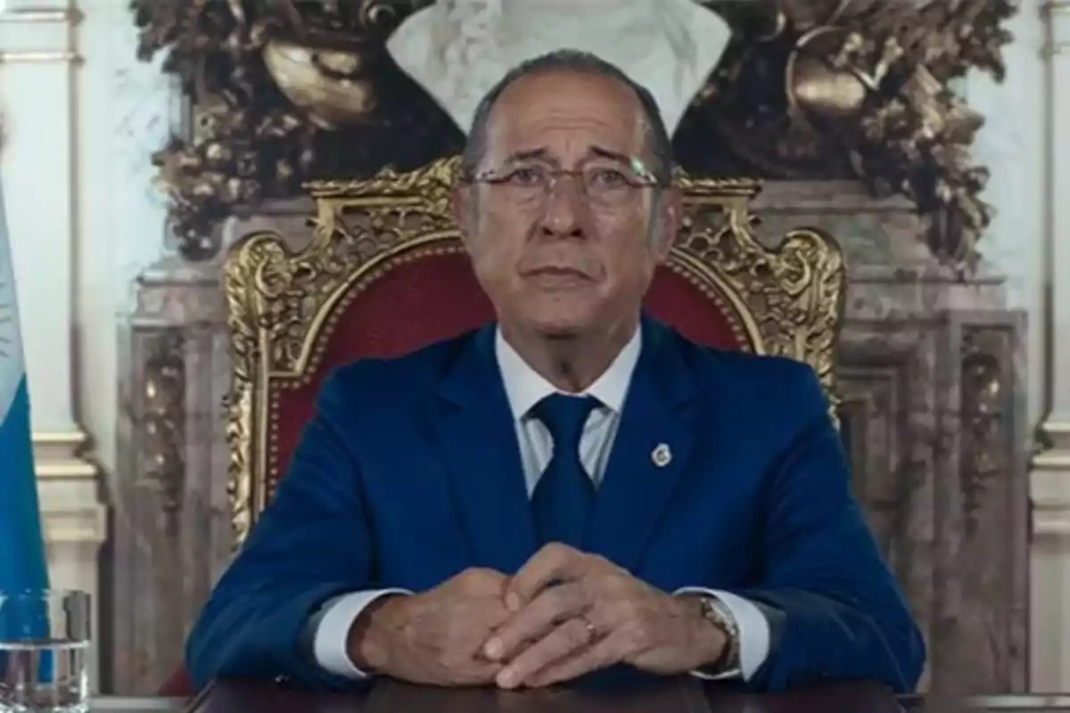 Man in a blue suit sitting on an elegant chair with a decorative background and a flag to the left Man in a blue suit sitting on an elegant chair with a decorative background and a flag to the left