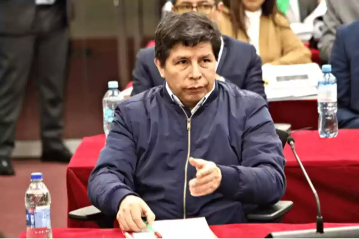 Hombre sentado en una sala de reuniones con mesas rojas y botellas de agua, sosteniendo un bolígrafo y mirando al frente