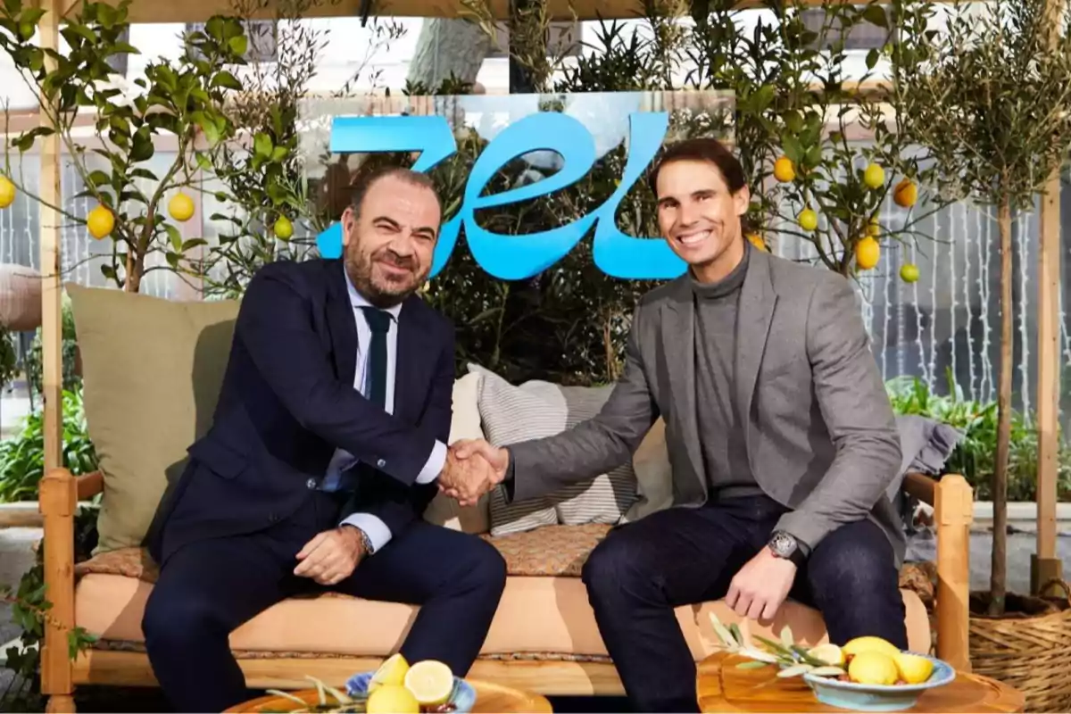 Dos hombres sonrientes se dan la mano sentados en un sofá rodeados de plantas y limones con un letrero azul de fondo