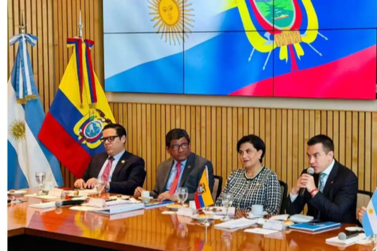 Cuatro personas sentadas en una mesa de reunión con banderas de Argentina y Ecuador, al fondo una pantalla muestra ambas banderas y hay documentos y micrófonos sobre la mesa Cuatro personas sentadas en una mesa de reunión con banderas de Argentina y Ecuador, al fondo una pantalla muestra ambas banderas y hay documentos y micrófonos sobre la mesa