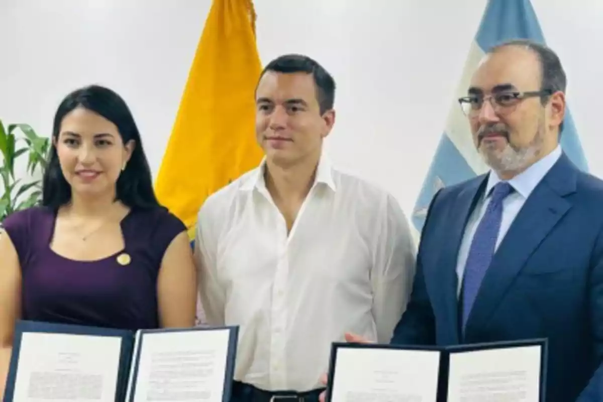 Tres personas posan para una foto formal, dos de ellas sostienen documentos y al fondo se observan dos banderas. Tres personas posan para una foto formal, dos de ellas sostienen documentos y al fondo se observan dos banderas.