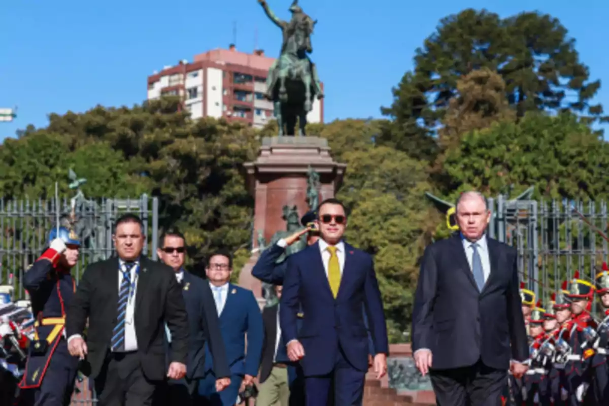 Grupo de hombres con trajes formales caminando frente a una estatua ecuestre y rodeados de guardias con uniformes militares en un entorno urbano con árboles y edificios al fondo