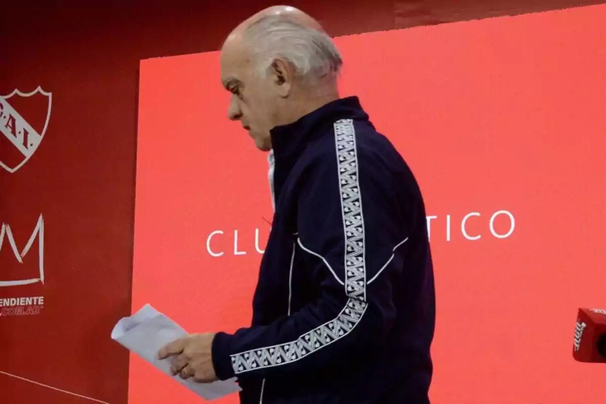 Man in profile wearing a blue sports jacket holding papers in front of a red background with the crest and name of a soccer club Man in profile wearing a blue sports jacket holding papers in front of a red background with the crest and name of a soccer club
