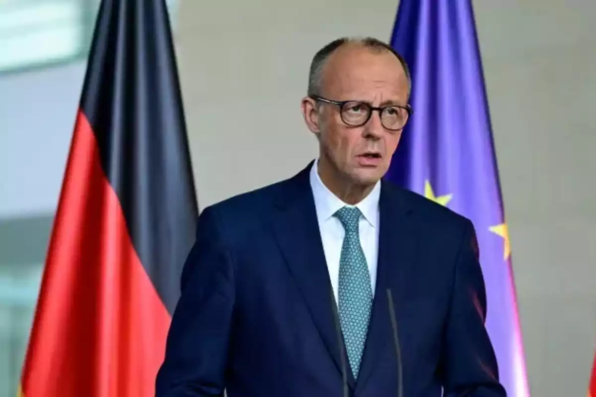 Man in suit and tie speaking in front of a microphone with German and European Union flags in the background
