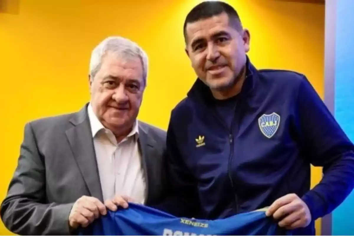 Dos hombres posan juntos frente a una pared amarilla, uno de ellos lleva un saco gris y el otro una campera azul con el escudo de Boca Juniors, ambos sostienen una camiseta azul. Dos hombres posan juntos frente a una pared amarilla, uno de ellos lleva un saco gris y el otro una campera azul con el escudo de Boca Juniors, ambos sostienen una camiseta azul.