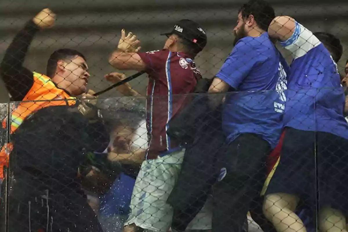 Un grupo de personas discute y se enfrenta físicamente en las gradas de un estadio detrás de una malla de protección Un grupo de personas discute y se enfrenta físicamente en las gradas de un estadio detrás de una malla de protección