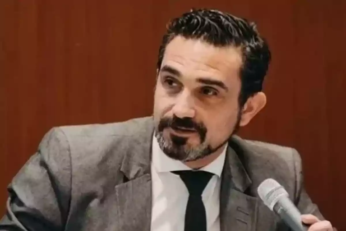 Hombre de cabello oscuro y barba con traje gris y corbata negra sosteniendo un micrófono frente a un fondo marrón