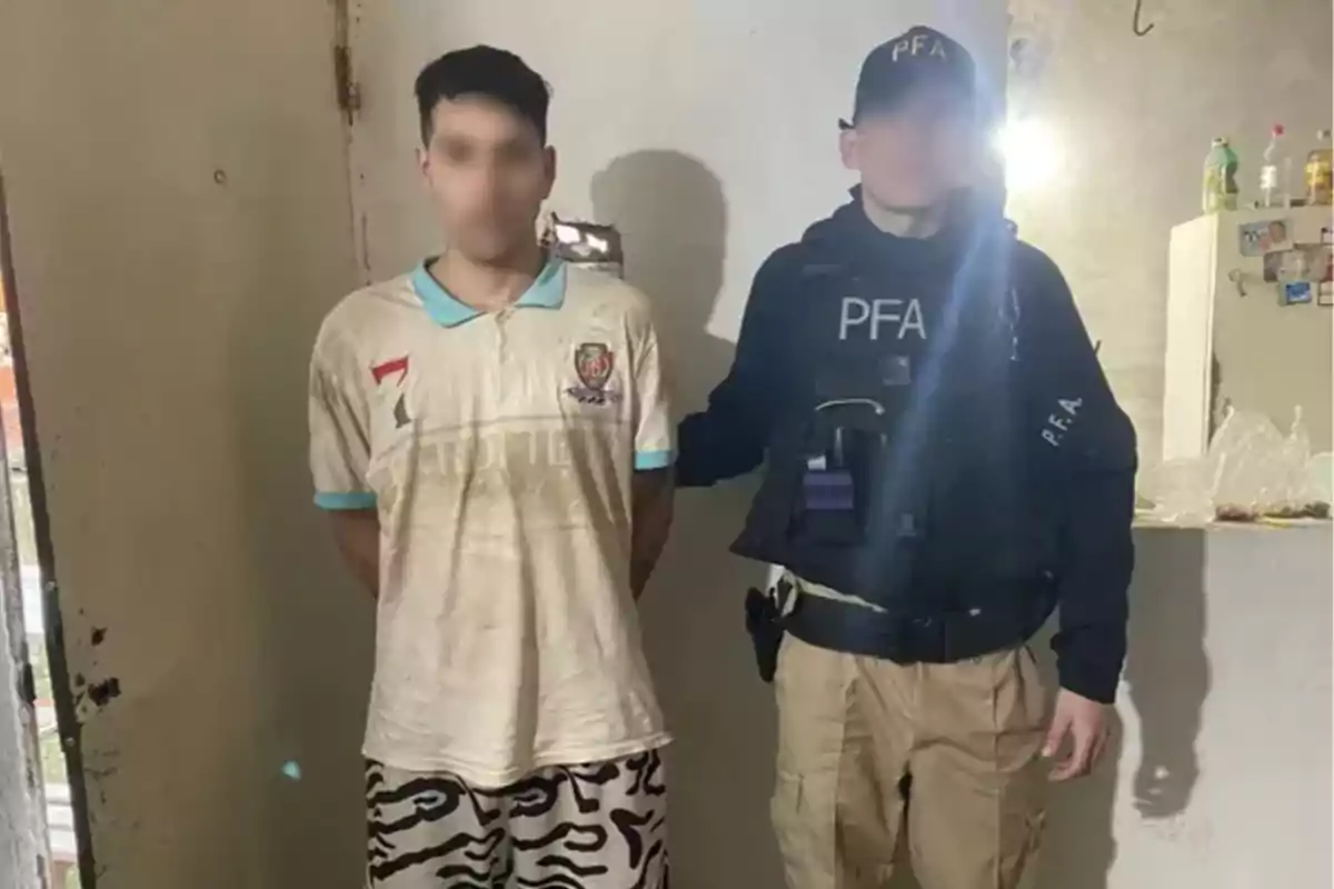 Un hombre con las manos hacia atrás es escoltado por un agente de la policía federal dentro de una vivienda.