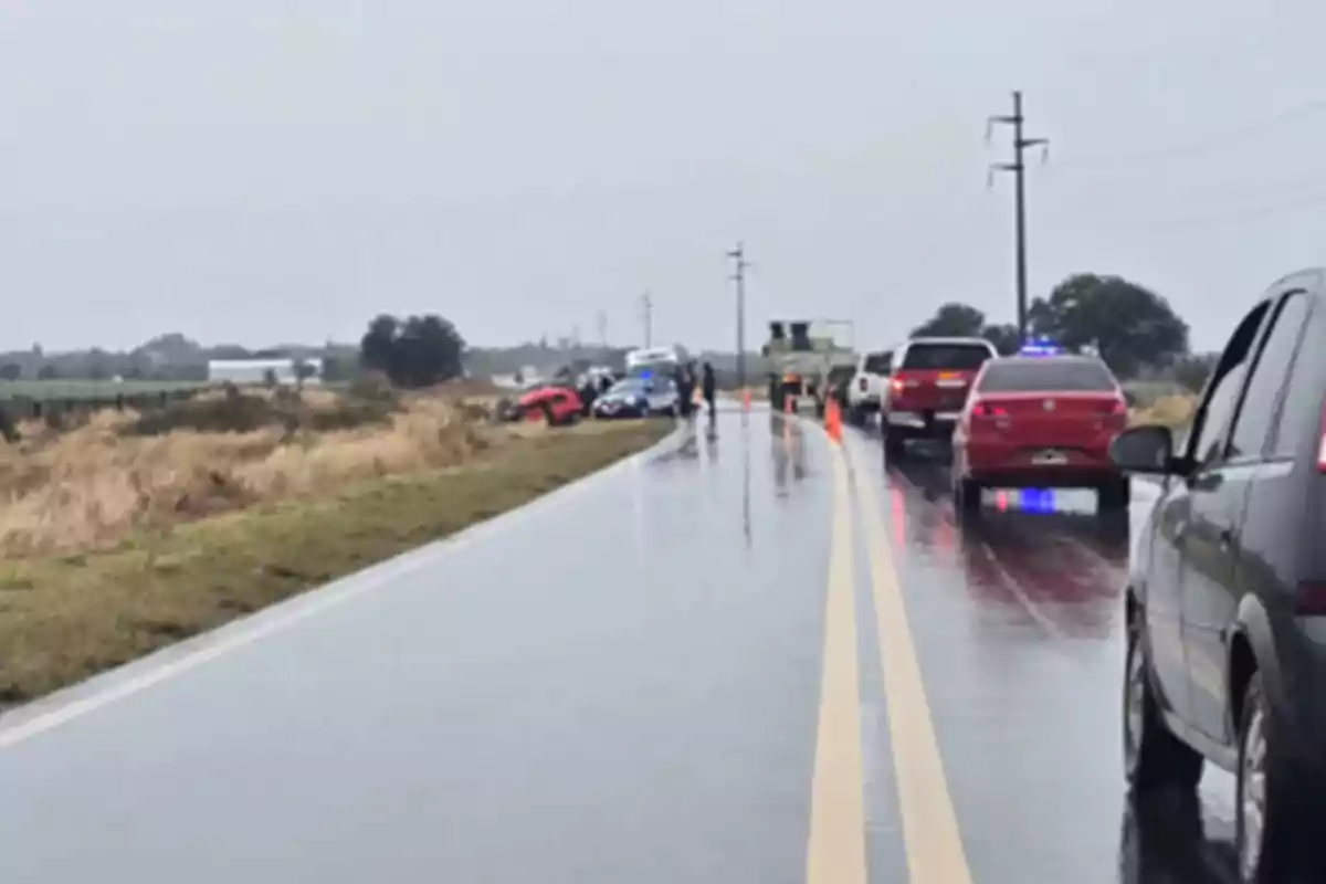 Several cars stopped on a rain-soaked road while a red car is off the road and police and emergency personnel are present. Several cars stopped on a rain-soaked road while a red car is off the road and police and emergency personnel are present.