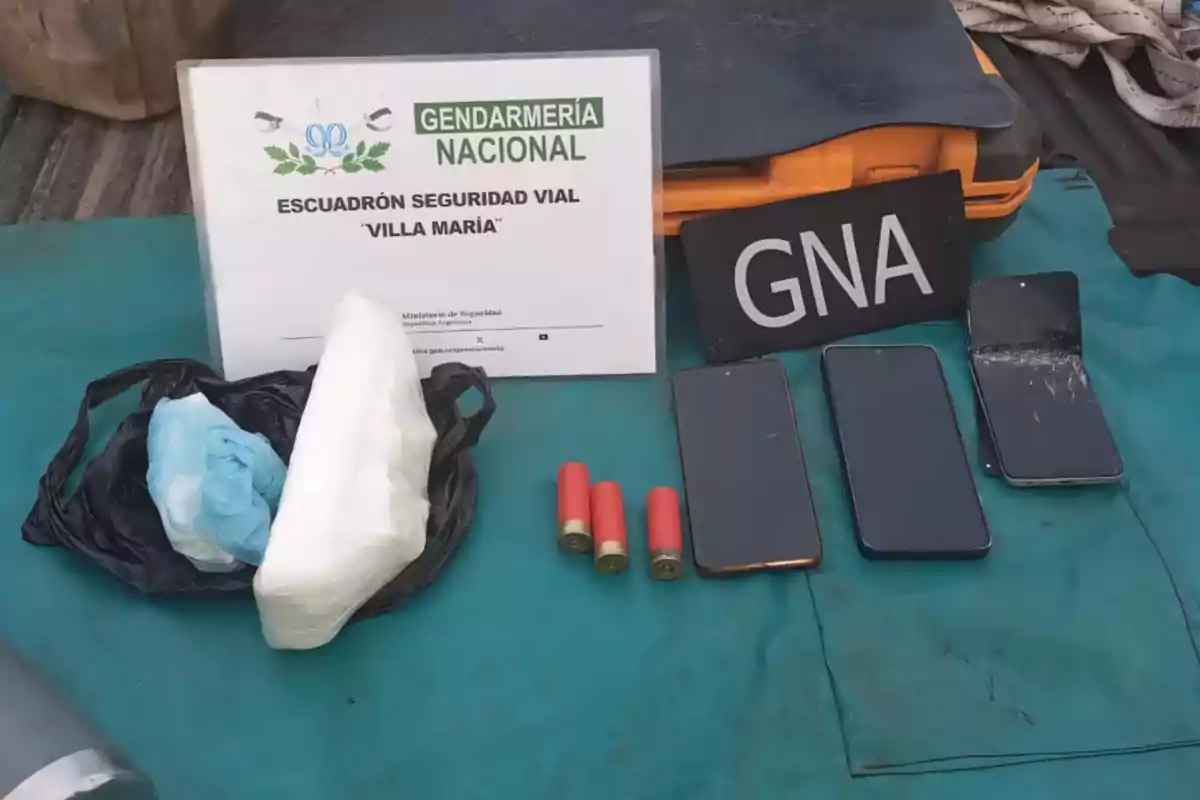 Sobre una superficie azul se observan tres teléfonos móviles, tres cartuchos de escopeta, un paquete envuelto en plástico, una bolsa negra, un cartel de la Gendarmería Nacional y una caja naranja Sobre una superficie azul se observan tres teléfonos móviles, tres cartuchos de escopeta, un paquete envuelto en plástico, una bolsa negra, un cartel de la Gendarmería Nacional y una caja naranja