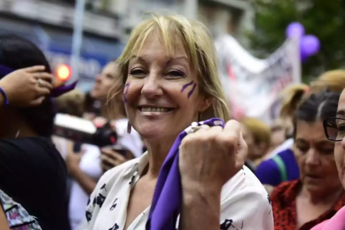 Mujer sonriente con pintura violeta en el rostro sostiene un pañuelo del mismo color durante una manifestación rodeada de otras personas Mujer sonriente con pintura violeta en el rostro sostiene un pañuelo del mismo color durante una manifestación rodeada de otras personas