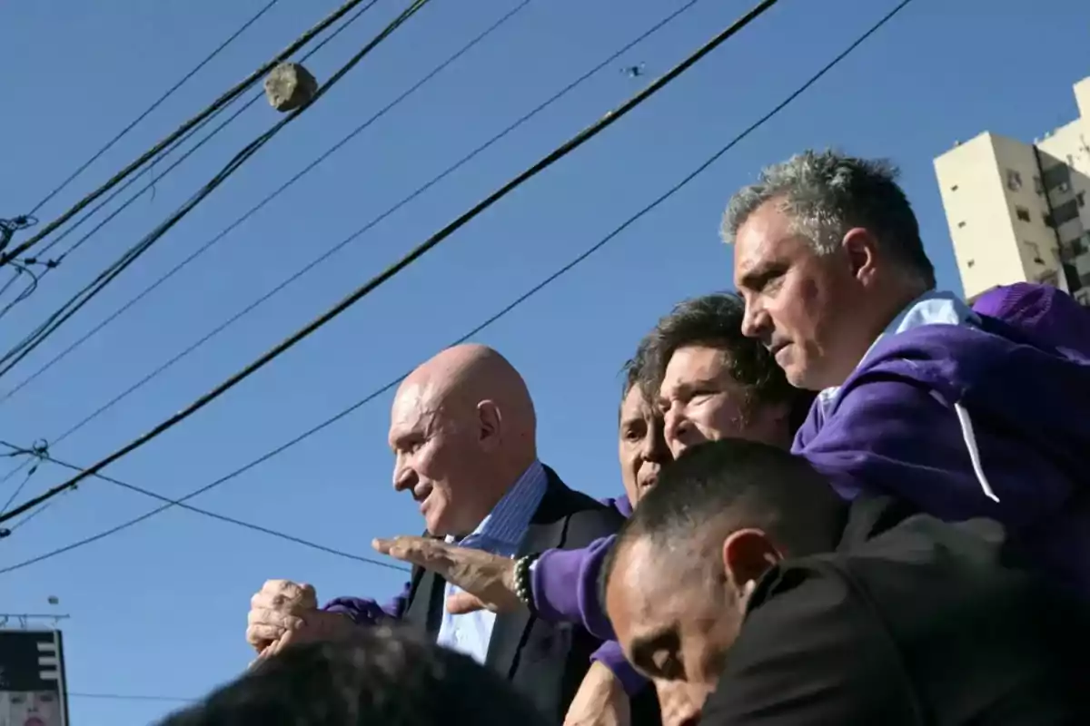 Group of men gathered outdoors under power lines with buildings in the background and a clear sky