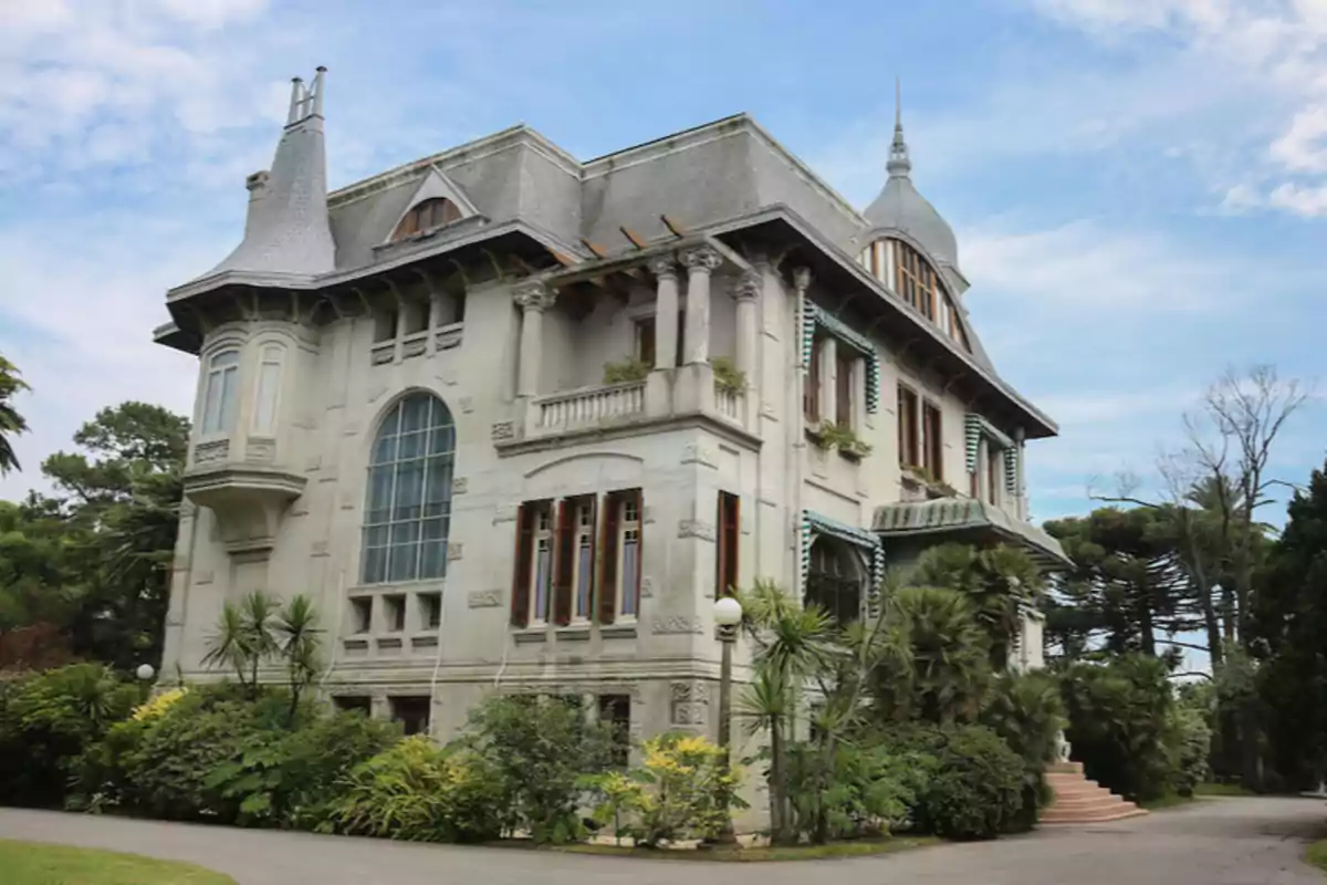 Old multi-story mansion with a sloped roof and large windows, surrounded by vegetation and trees under a blue sky Old multi-story mansion with a sloped roof and large windows, surrounded by vegetation and trees under a blue sky
