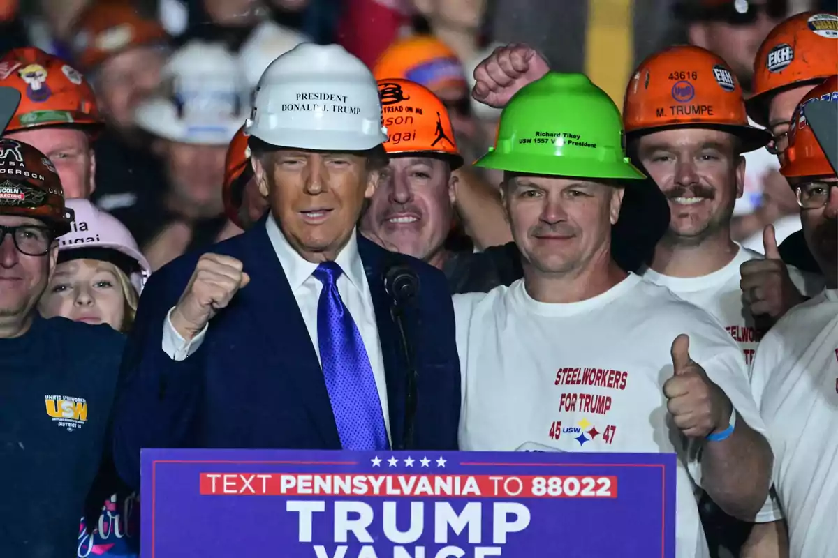 Donald Trump con casco blanco rodeado de trabajadores con cascos de colores en un evento de campaña, algunos levantando el pulgar y otros con el puño en alto