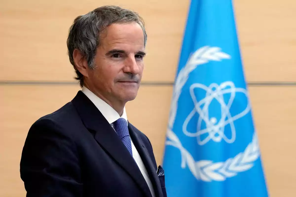 Gray-haired man in a dark suit and blue tie standing in front of a blue flag with a white atomic symbol