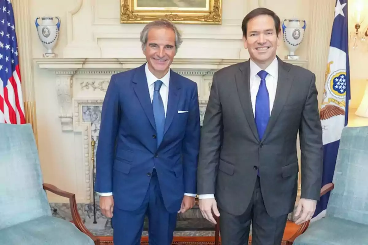 Two men in suits pose smiling in an elegant room with flags and a fireplace in the background. Two men in suits pose smiling in an elegant room with flags and a fireplace in the background.