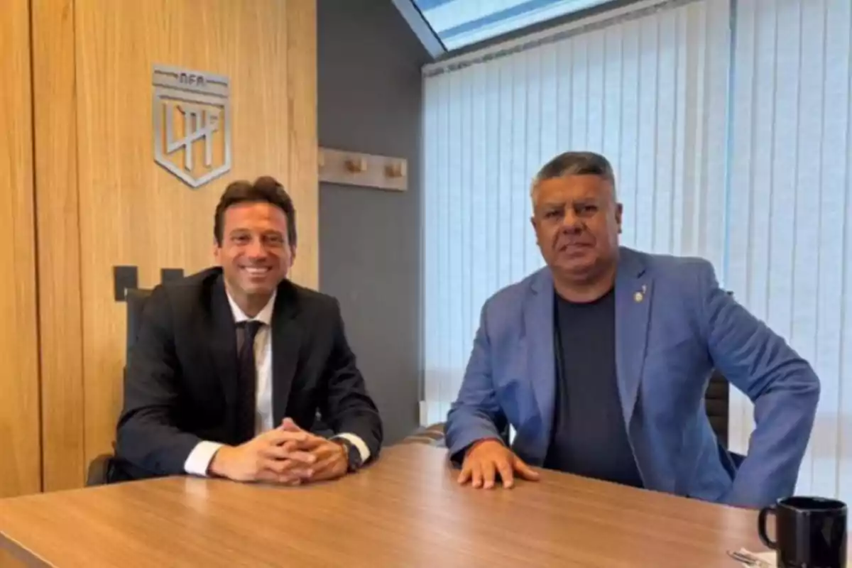 Two men sitting at an office table, both wearing jackets, one smiling and the other with a serious expression; in the background, there is a wooden wall with a logo and a window with vertical blinds. Two men sitting at an office table, both wearing jackets, one smiling and the other with a serious expression; in the background, there is a wooden wall with a logo and a window with vertical blinds.