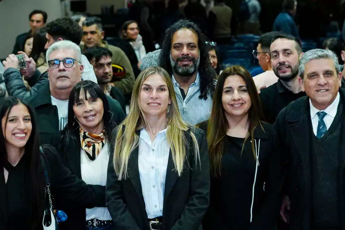 Grupo de personas posando y sonriendo en un evento social o profesional, con otras personas al fondo Grupo de personas posando y sonriendo en un evento social o profesional, con otras personas al fondo