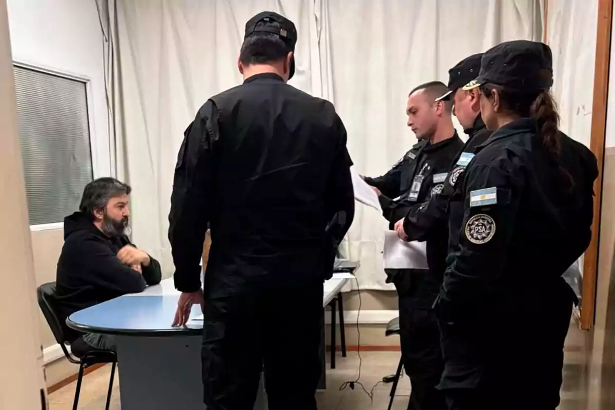 A man sitting at a table is surrounded by four security officers who are checking documents in an office.