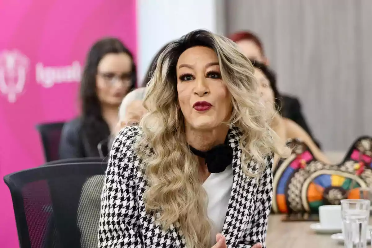 Person with long blonde hair, wearing a plaid jacket, sitting at a table during a meeting; in the background, other people and a pink wall can be seen. Person with long blonde hair, wearing a plaid jacket, sitting at a table during a meeting; in the background, other people and a pink wall can be seen.