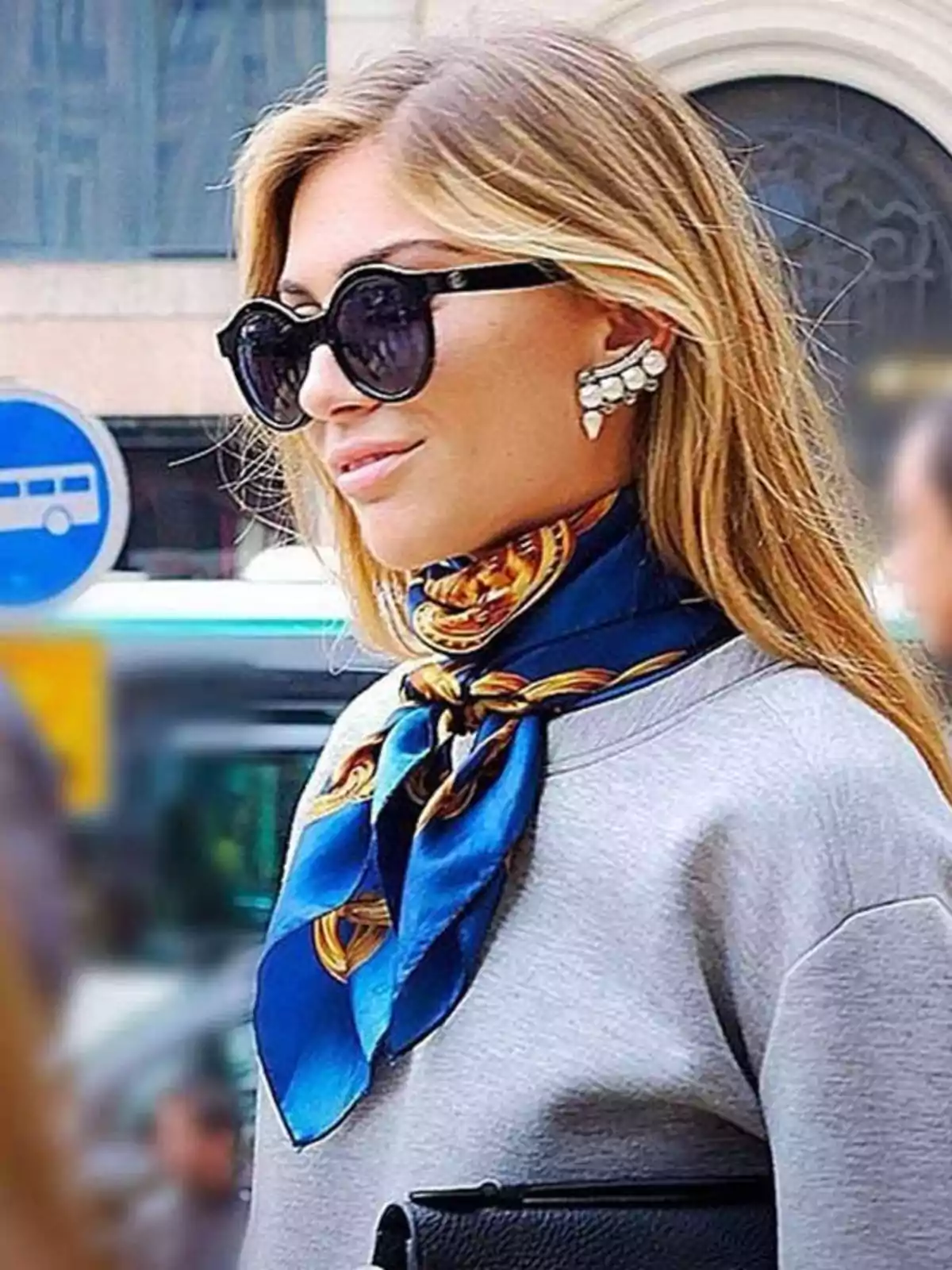 Blonde woman with large sunglasses, a blue scarf around her neck, striking earrings, and wearing a gray sweater in an urban setting Blonde woman with large sunglasses, a blue scarf around her neck, striking earrings, and wearing a gray sweater in an urban setting