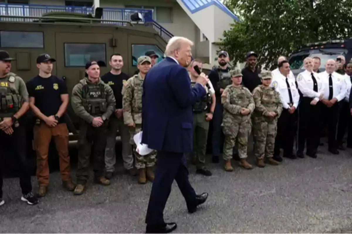 A man in a blue suit holds a microphone and addresses a group of police officers and soldiers lined up next to an armored vehicle in an urban setting.