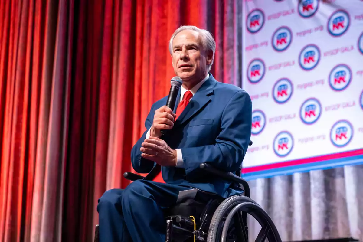 Gray-haired man in a wheelchair wearing a blue suit and red tie while holding a microphone on a stage with red curtains and a background with circular logos