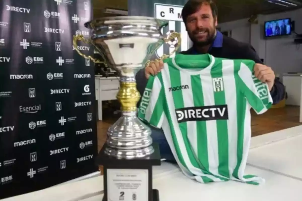 Hombre sonriente sostiene una camiseta de fútbol verde y blanca junto a un gran trofeo plateado en una oficina Hombre sonriente sostiene una camiseta de fútbol verde y blanca junto a un gran trofeo plateado en una oficina