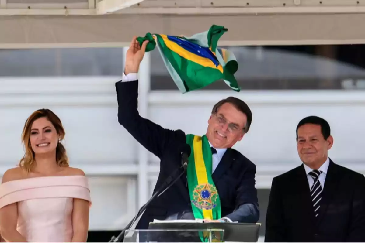 Un hombre con banda presidencial sostiene una bandera de Brasil mientras sonríe, acompañado por una mujer con vestido claro y otro hombre con traje oscuro.